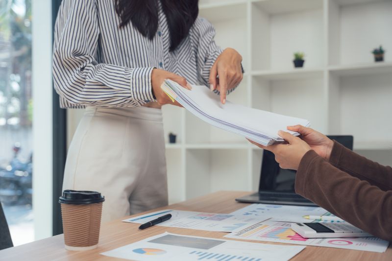 An employee handing a document across a desk to another person in a professional office, with paperwork and a coffee cup visible on the desk