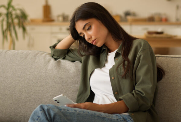 Woman sitting on couch looking at her phone with a worried expression after being let go from her job