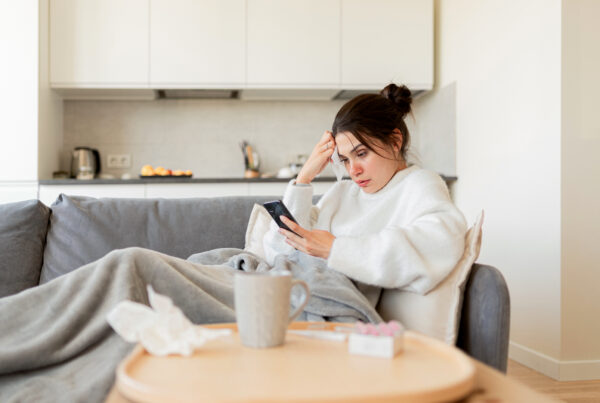 Worried woman sitting on her couch after being fired for calling in sick, looking at her phone while wrapped in a blanket with medicine nearby