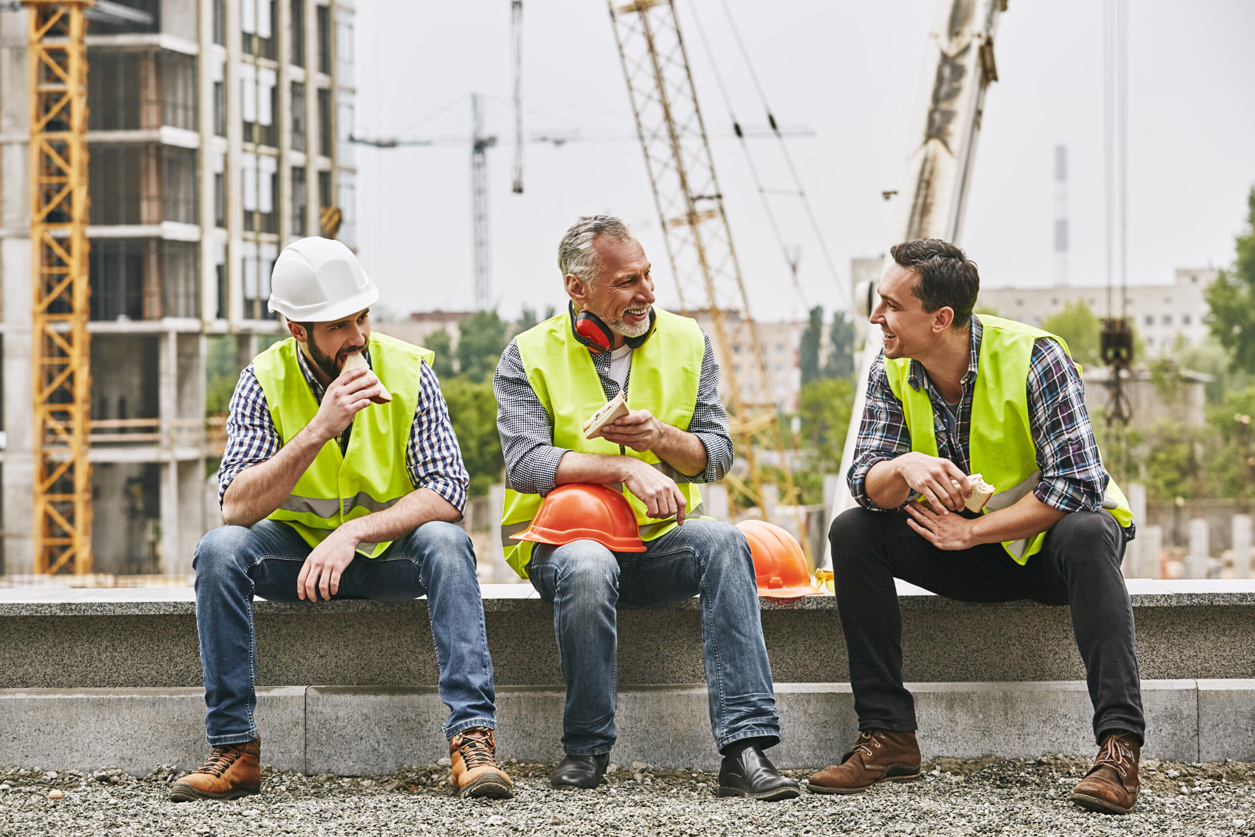 Three male construction workers in yellow safety vests sit on a ledge at an active construction site eating lunch and talking, illustrating California employees' right to uninterrupted meal breaks.