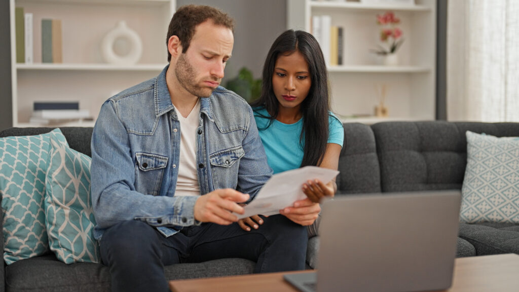 A man and woman sitting on a couch together reviewing a document, with a laptop open on the coffee table in front of them
