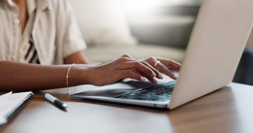 Close-up of a woman's hands typing on a laptop at a desk with a notebook and pen beside her documenting employment history after receiving a PIP