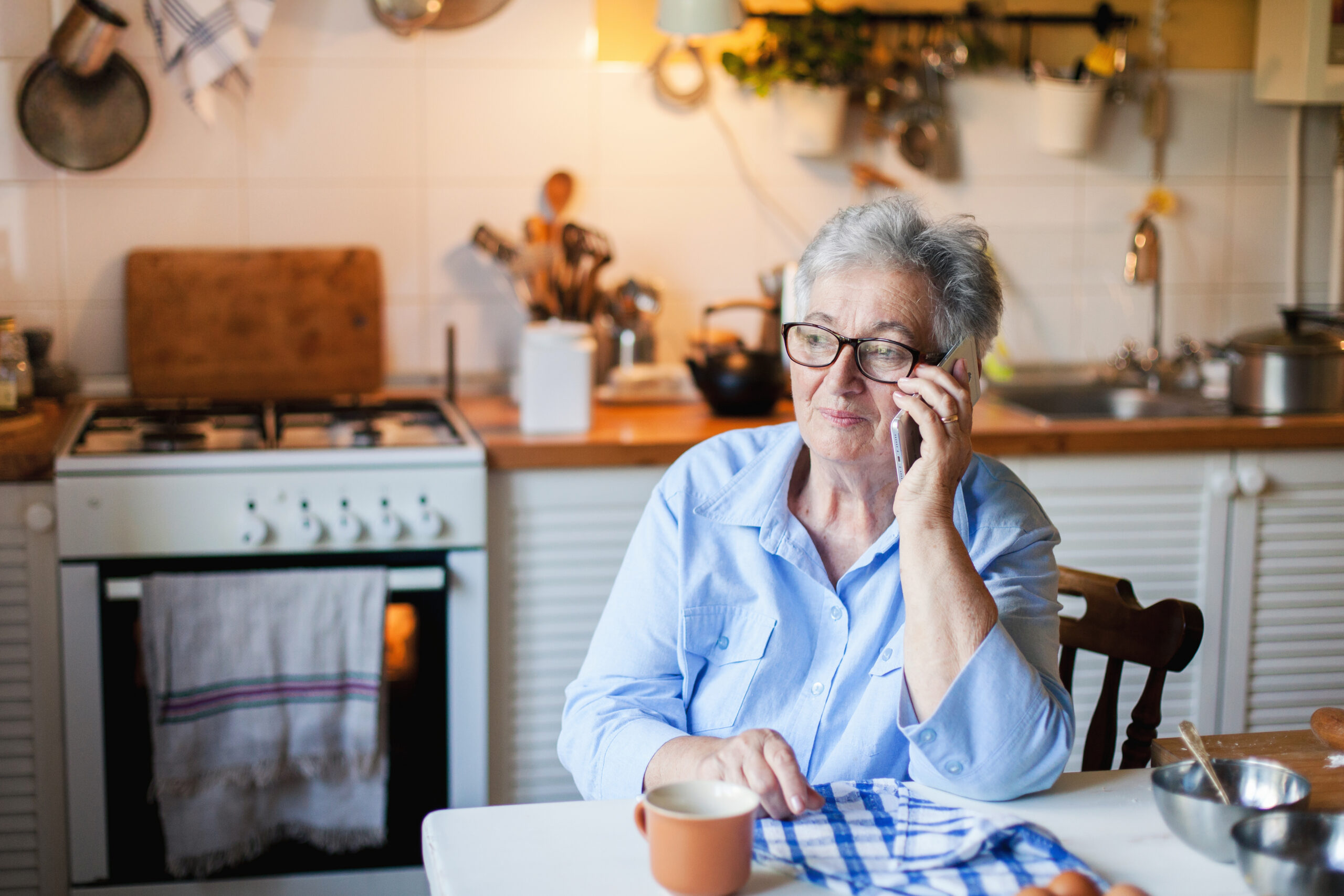 An older woman with gray hair and glasses sits at her kitchen table making a phone call with a focused expression, researching her options after a wage dispute with her employer.