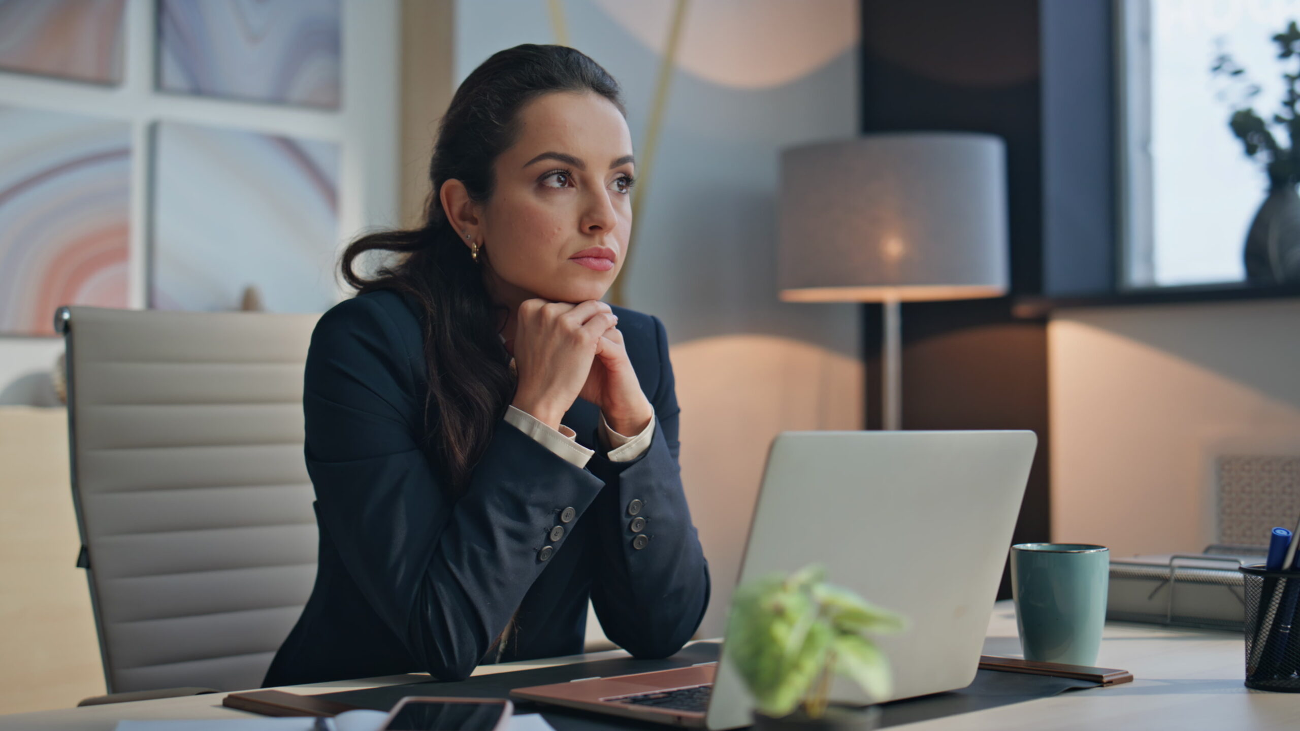A professional woman sits at her office desk with her hands clasped, looking thoughtfully off to the side with a laptop open in front of her, thinking about the sexual harassment at work she deals with.