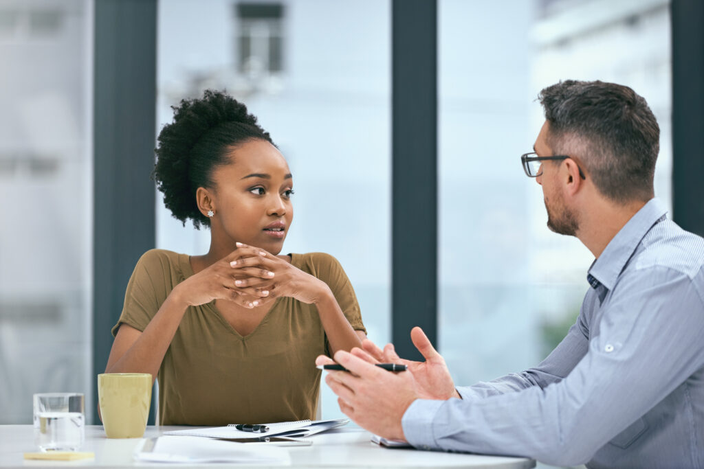 A woman listening attentively as a man explains the wrongful termination statute of limitations across a table in a bright office setting, with a notepad and coffee mug nearby