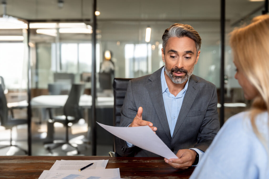 An attorney in a grey suit reviews a severance agreement document with a client across a desk in a modern office setting.