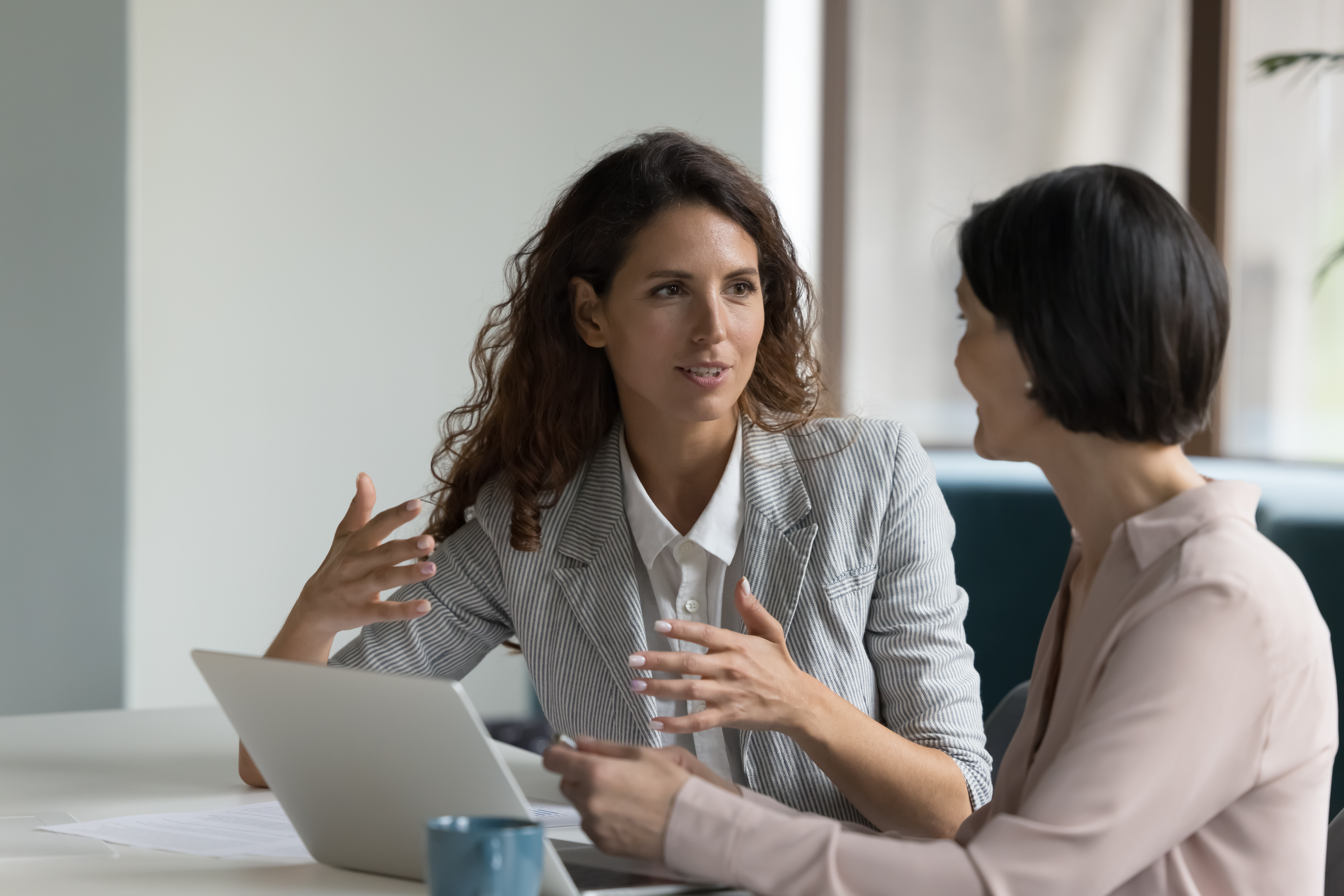 Two professional women sit together at a table in a modern office, one gesturing while explaining something and the other listening closely