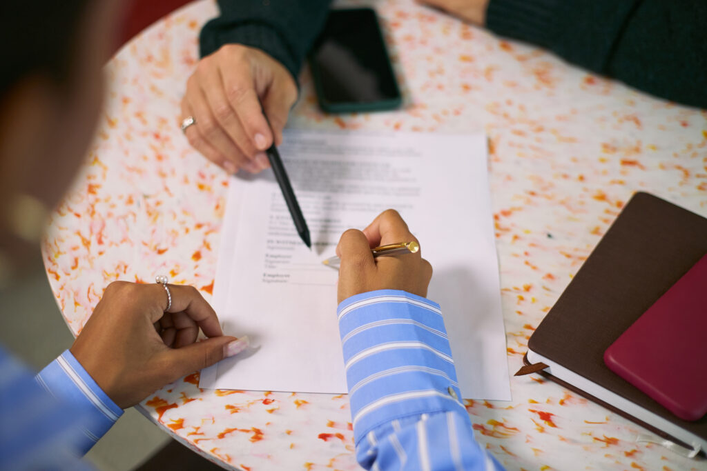 Overhead close-up of two people reviewing a contract with visible signature lines at a table, one person pointing with a pen before signing.