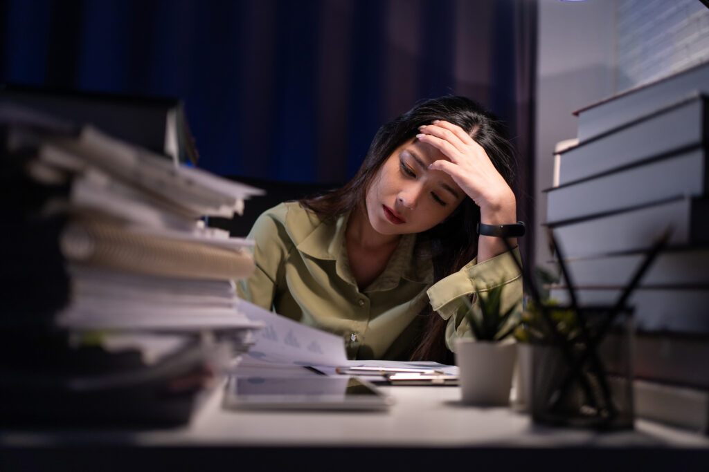 Employee exhausted at desk late at night surrounded by stacks of paperwork while working unpaid overtime