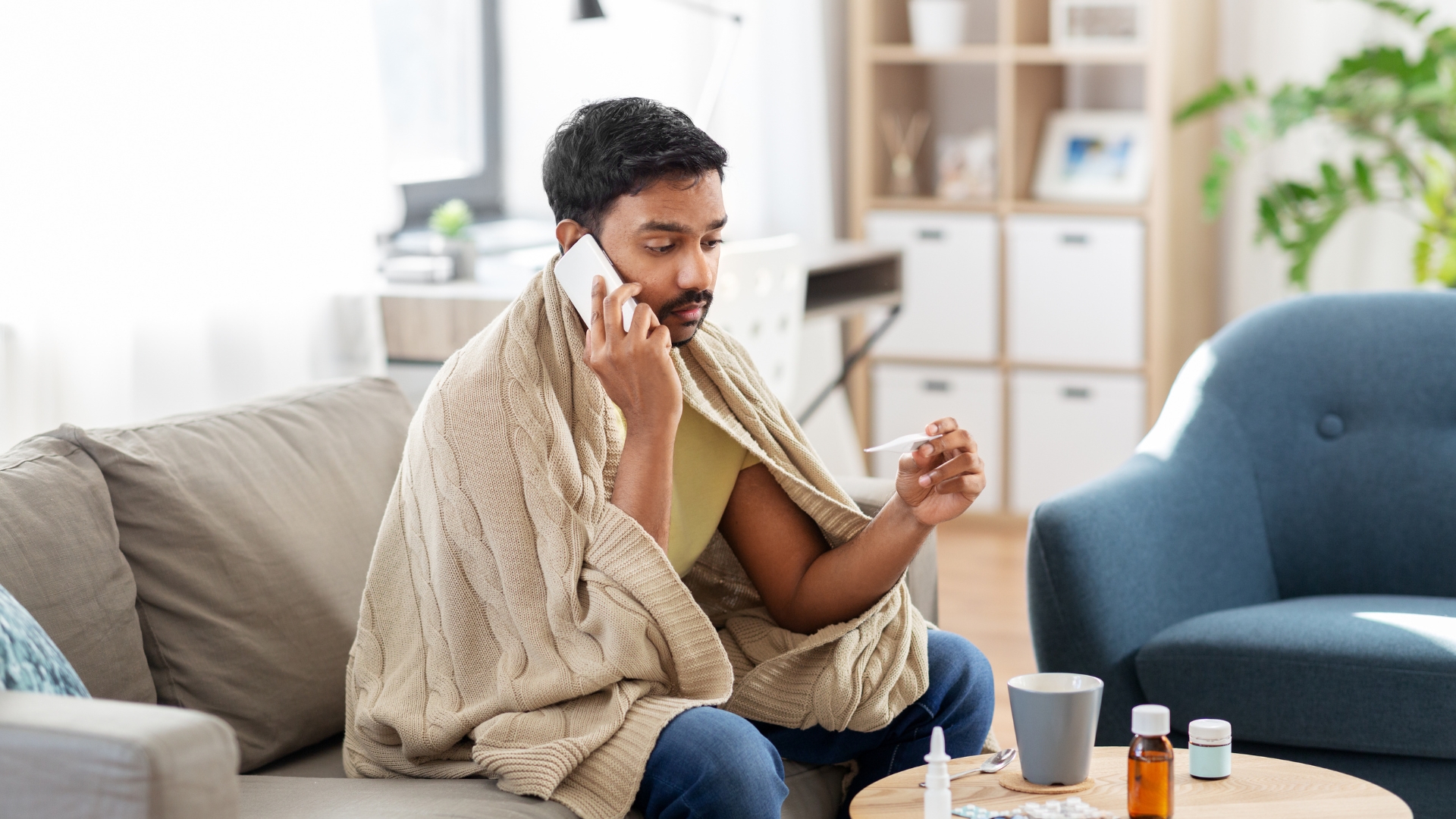 Sick man calling in sick from work while sitting on his couch at home, wrapped in a blanket and holding a thermometer with cold medicine on the table