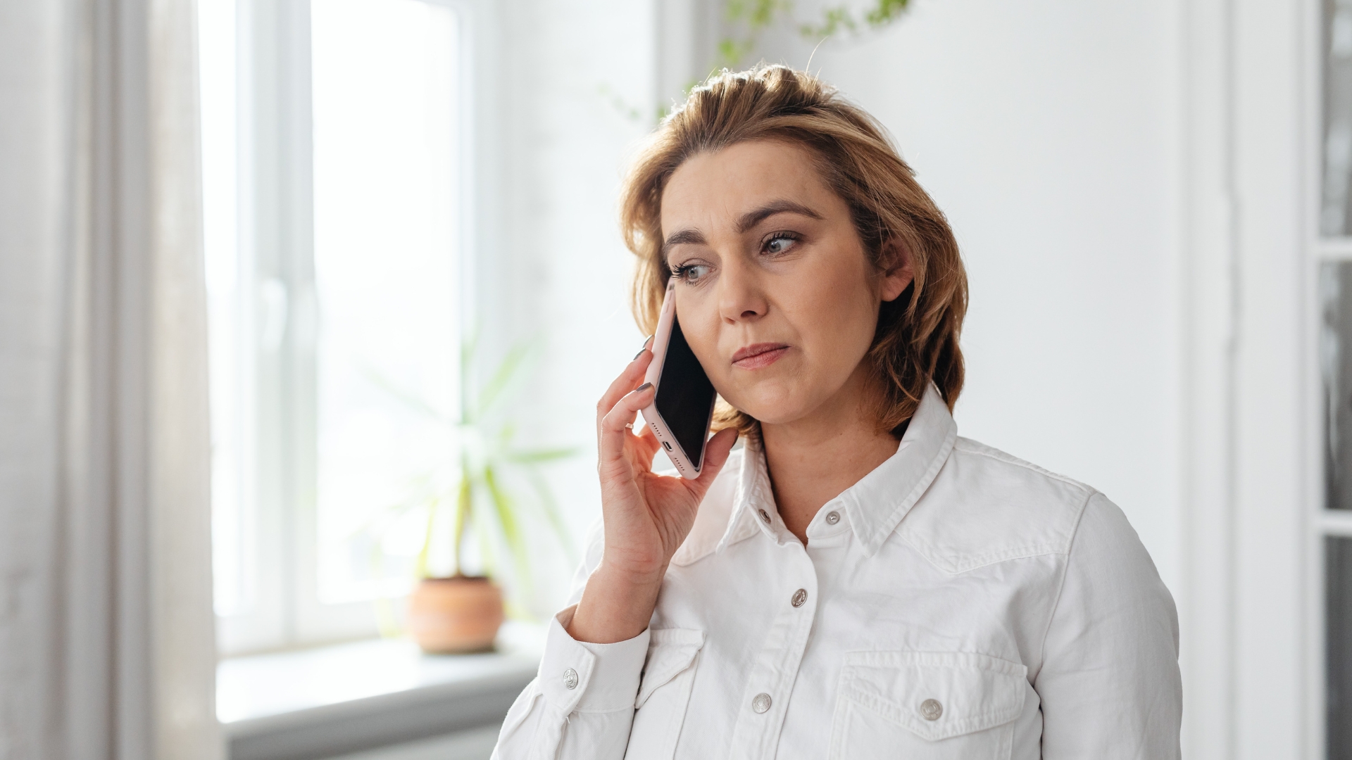 A woman in a white jacket taking a serious phone call near a bright window with a plant in the background, looking thoughtful and focused