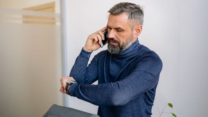 A man in a navy turtleneck standing with his arms crossed, talking on a phone with a serious expression in a modern interior setting