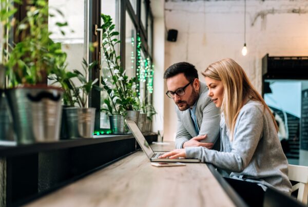 Two people leaning over a laptop together at a window-side counter surrounded by plants, reviewing information in a bright modern space