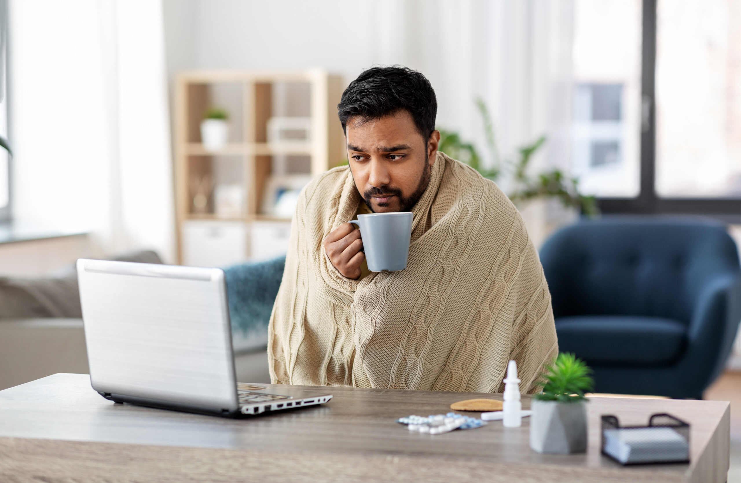 Asian man wrapped in a blanket holding a tea mug while looking at a laptop at home, surrounded by medication on the desk, representing an employee dealing with a serious health condition who may qualify for FMLA or CFRA protected medical leave in California