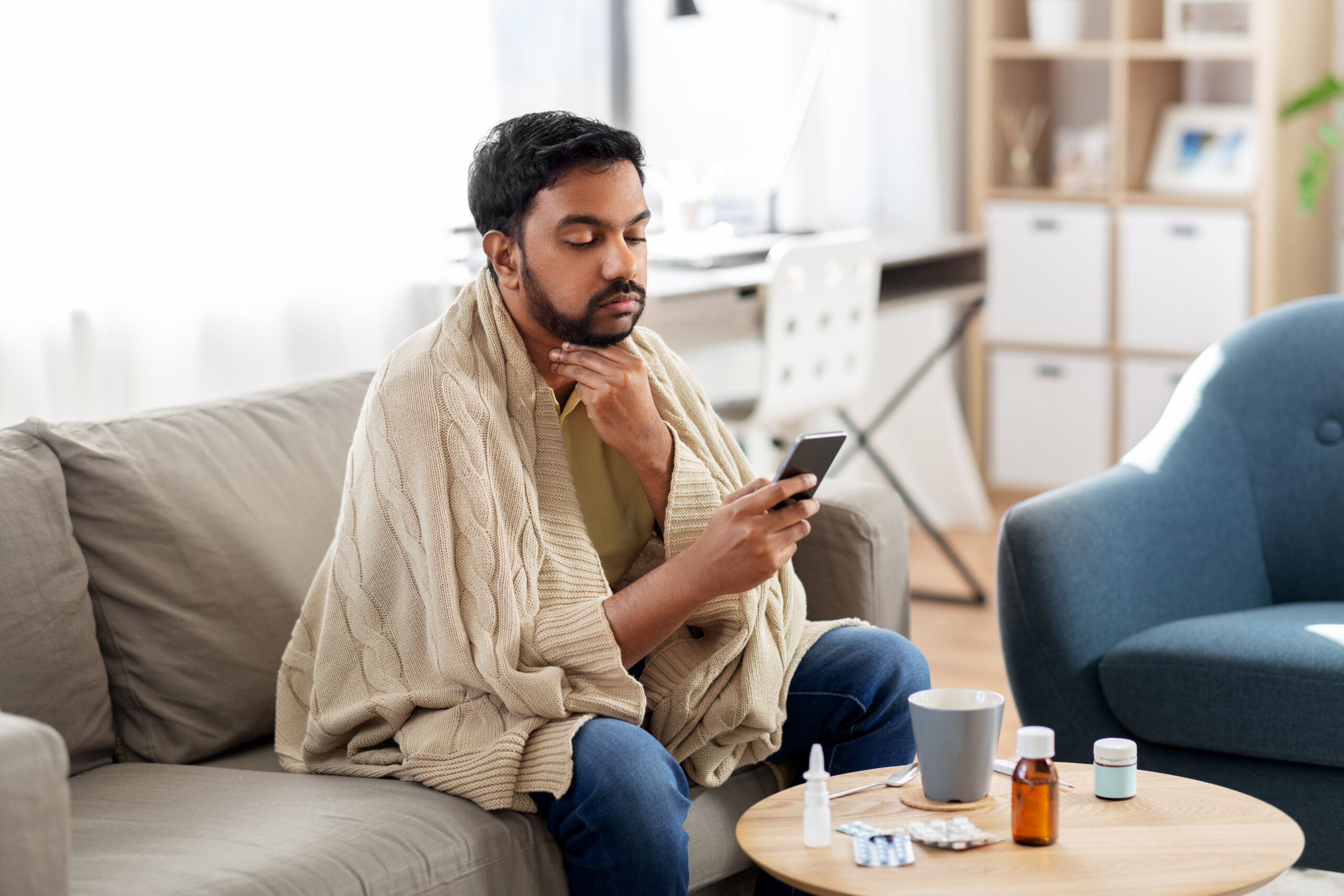 man wrapped in a blanket sitting on a couch looking at his phone with a thoughtful expression, medication on the coffee table in front of him, representing a California employee on medical leave researching their FMLA and CFRA rights