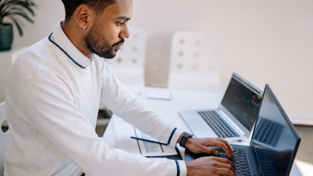 A man in a white sweater working focused at a laptop in a bright office, with a second laptop and handwritten notes on the desk beside him