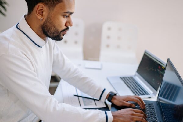A man in a white sweater working focused at a laptop in a bright office, with a second laptop and handwritten notes on the desk beside him