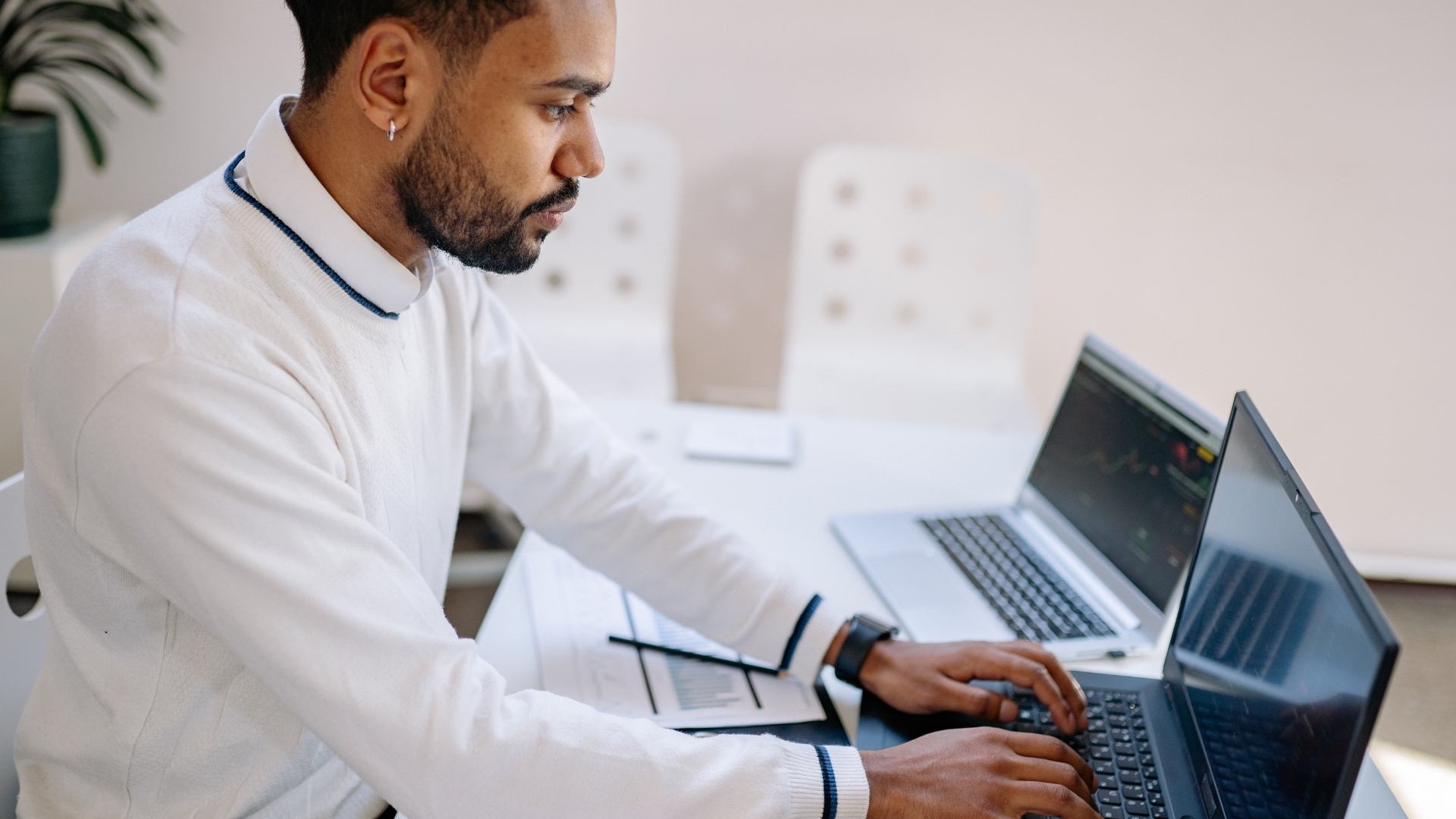 A man in a white sweater working focused at a laptop in a bright office, with a second laptop and handwritten notes on the desk beside him