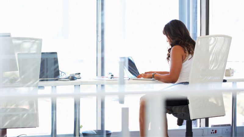 Woman sitting alone at a large office desk in a bright modern workspace, representing workplace isolation as a potential sign of a hostile work environment under FEHA California