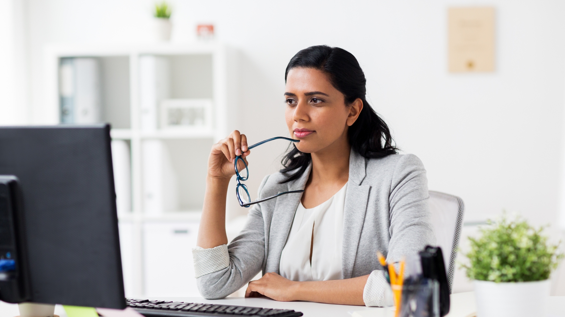 A professional woman at her office desk holds her glasses and looks at her screen with a thoughtful expression, considering whether her workplace qualifies as a hostile work environment