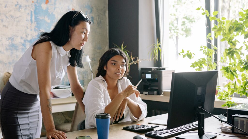 A supervisor leans over a seated employee reviewing work on a computer screen, a workplace dynamic that can become a warning sign of a hostile work environment