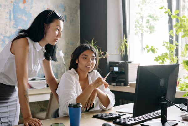 A supervisor leans over a seated employee reviewing work on a computer screen, a workplace dynamic that can become a warning sign of a hostile work environment