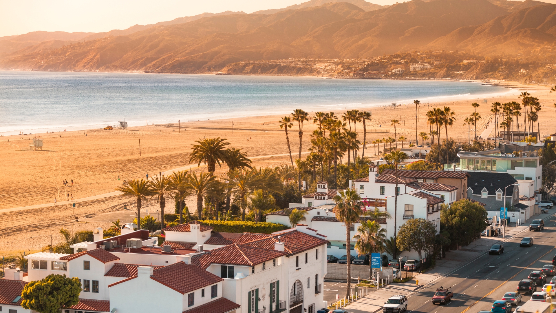 Aerial view of a California coastal town at sunset, with sandy beach, palm-lined road, and Spanish-style homes