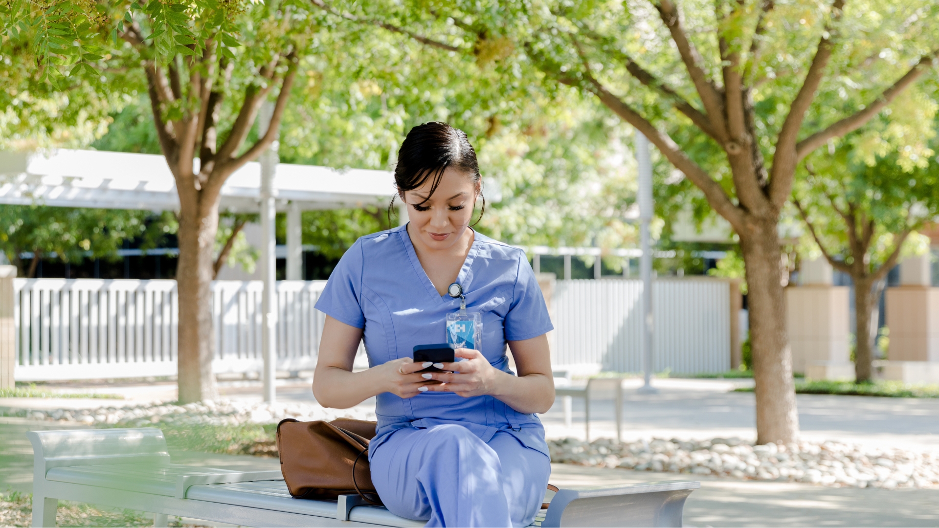 A nurse in scrubs sits outside on a bench checking her phone during a break from a California 12-hour shift