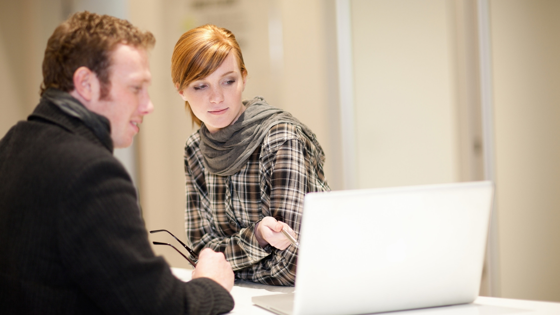 Two colleagues discuss a workplace decision while reviewing a laptop screen together in an office.