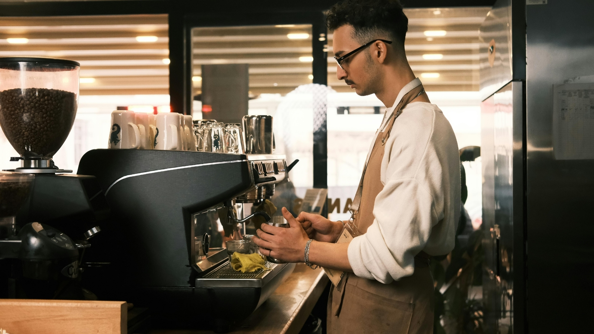 Barista in a tan apron steaming milk at an espresso machine in a warm cafe