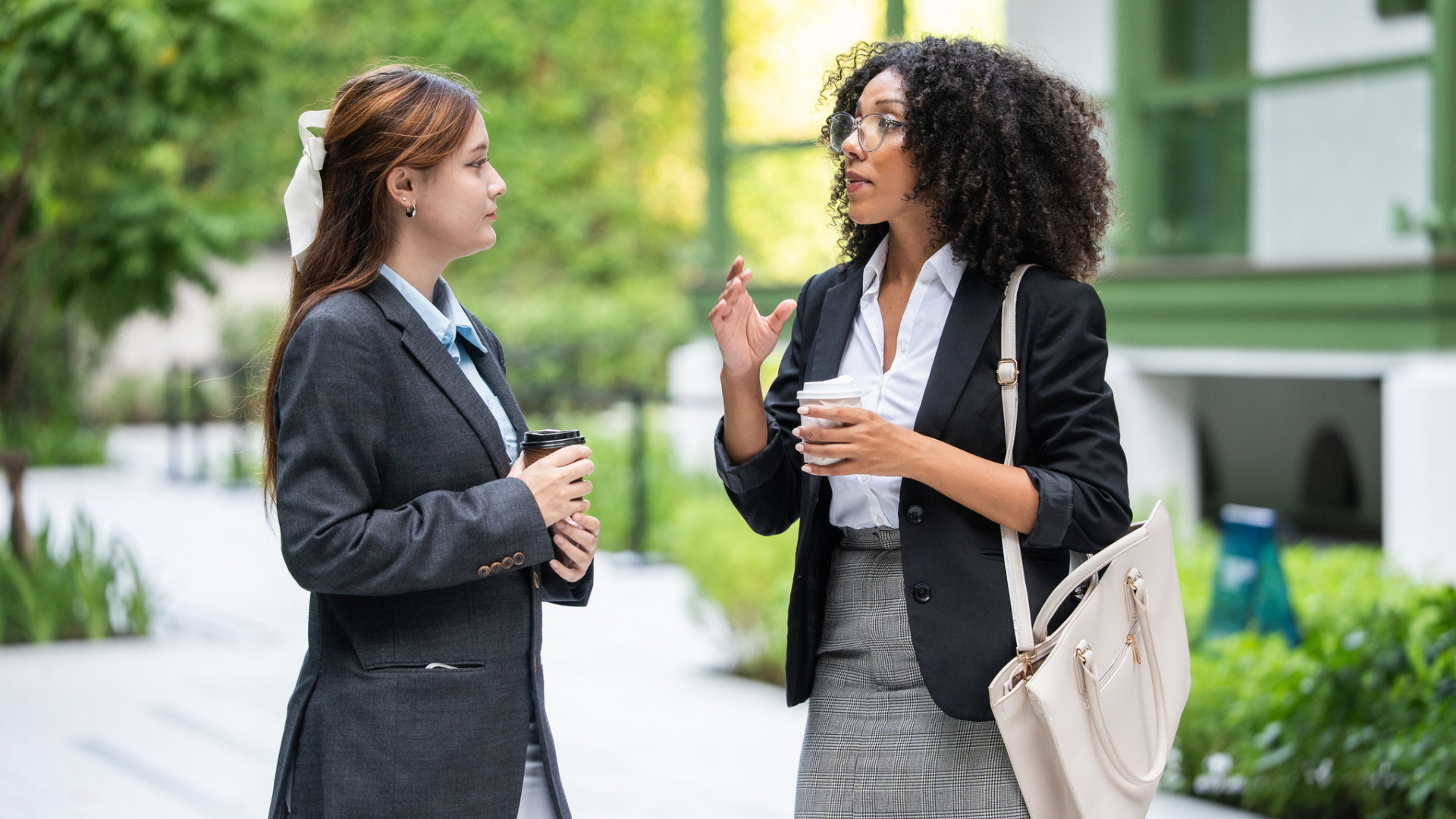 Two coworkers talking over coffee outside an office, one listening as the other explains a problem