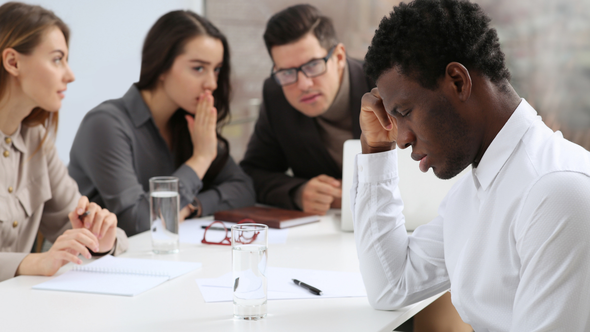 Employee sitting apart at a conference table while coworkers whisper during a workplace meeting
