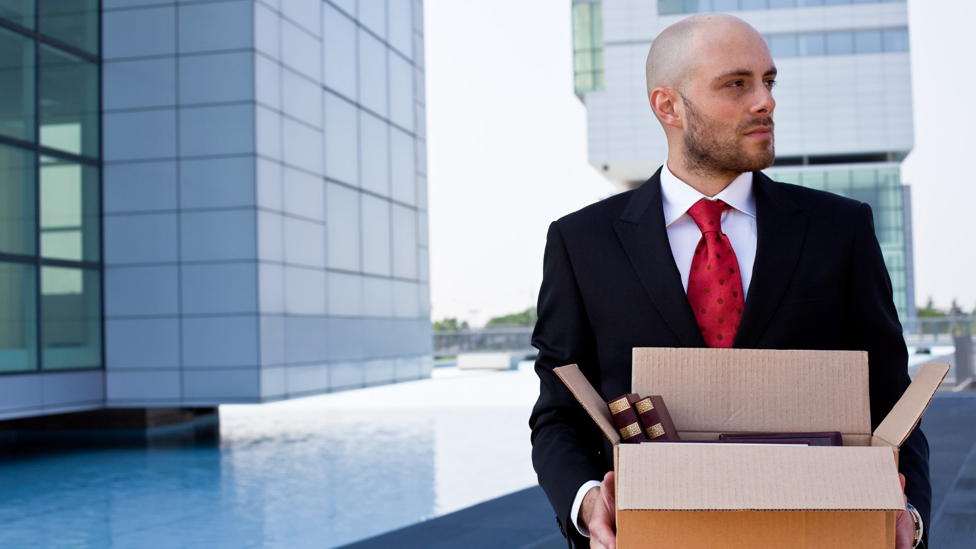 Man in a suit carries a box of belongings while walking away from a modern office building.