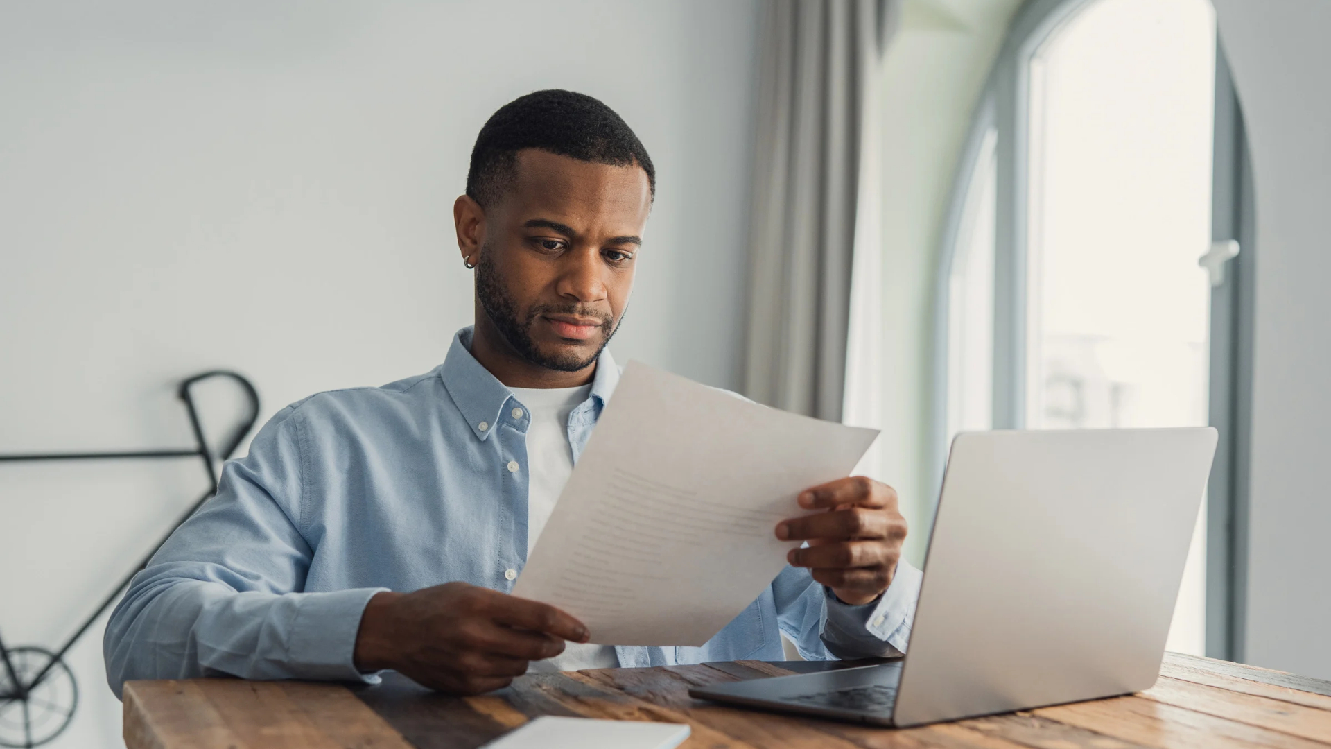 Employee reviewing a printed agreement at a sunlit table with a laptop open beside him