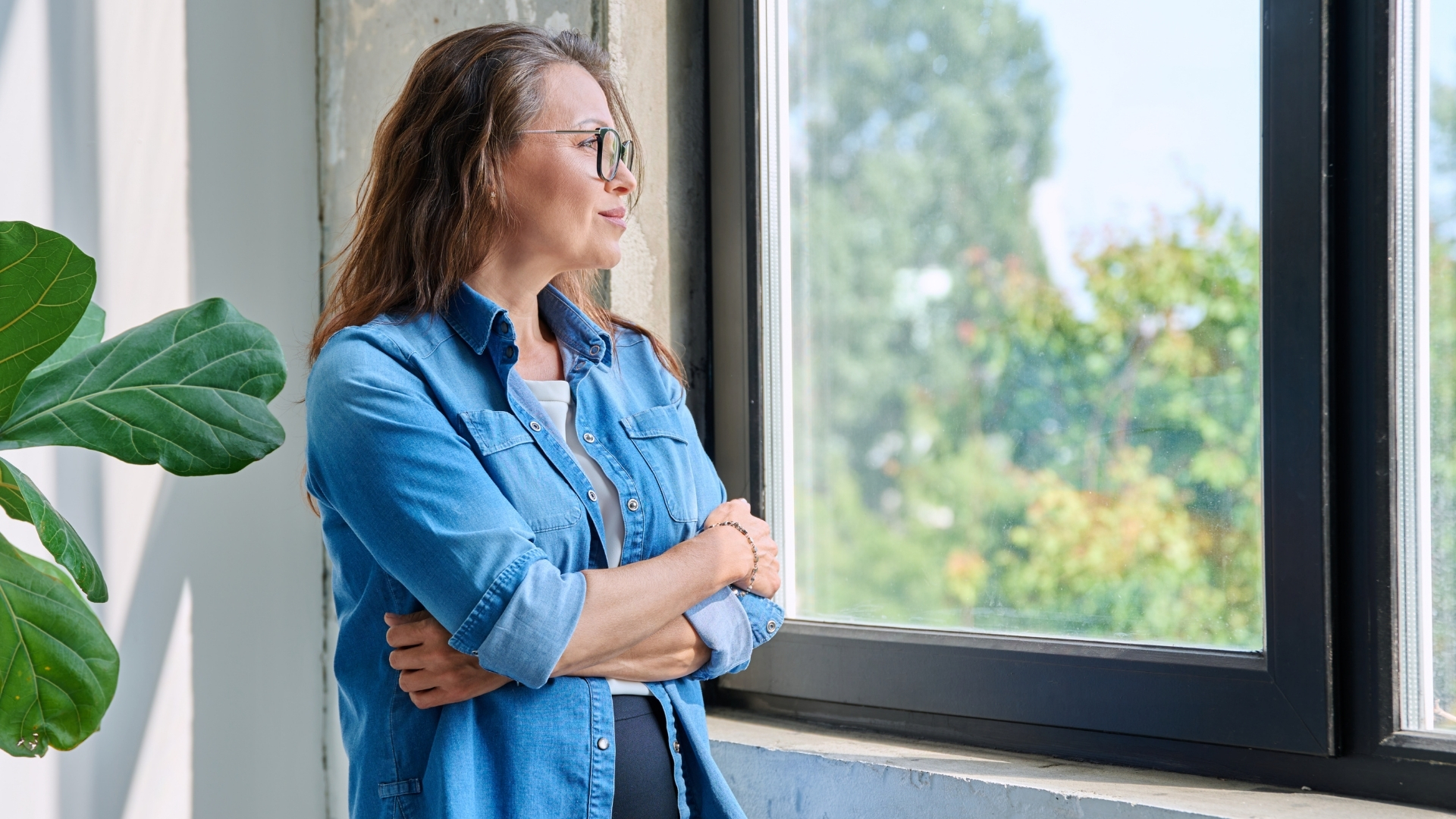 Woman in a denim shirt stands by a window with arms crossed, thinking quietly after leaving her job.