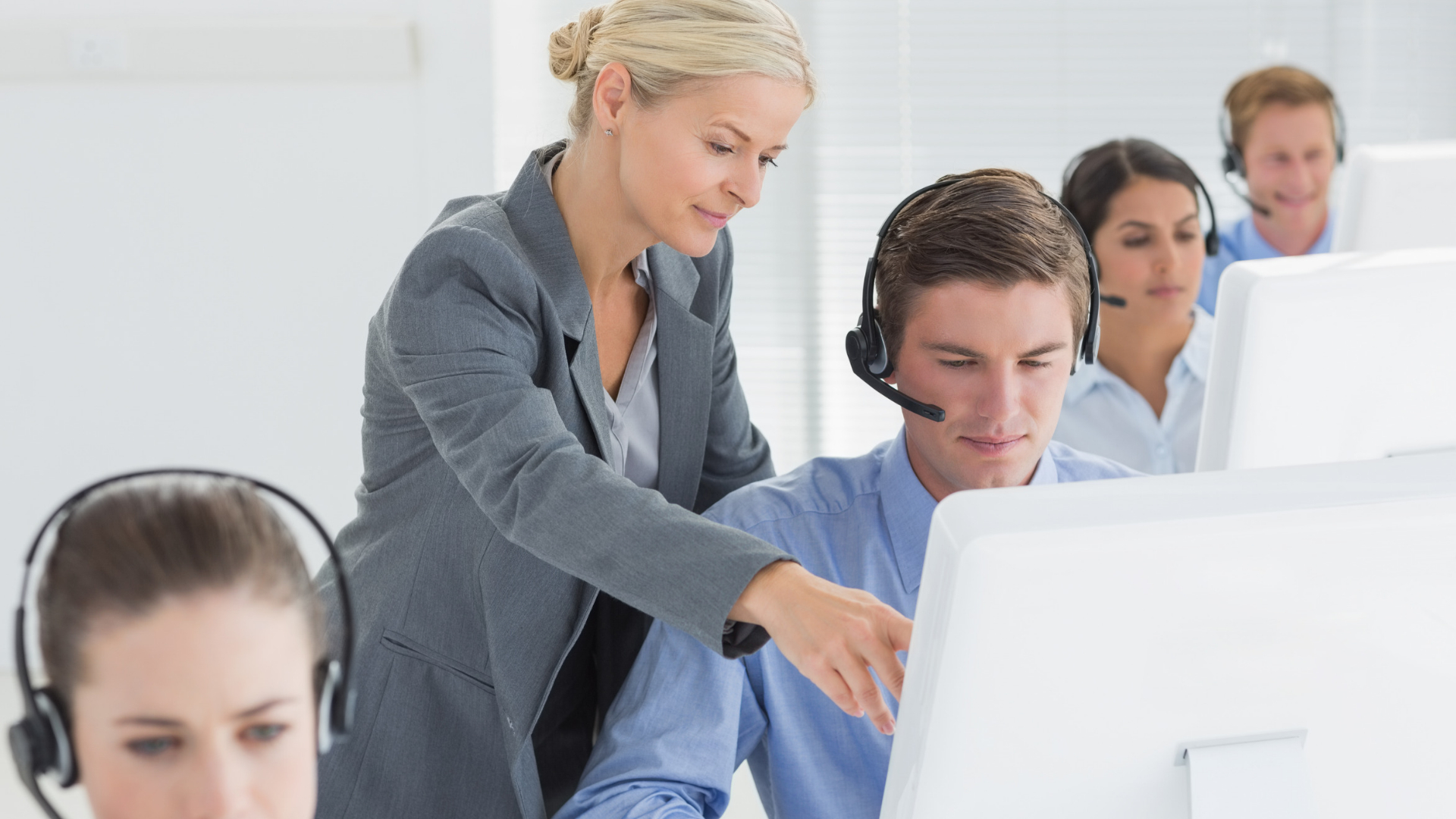 A manager pointing at a computer screen while a seated employee wearing a headset looks on, with coworkers nearby.