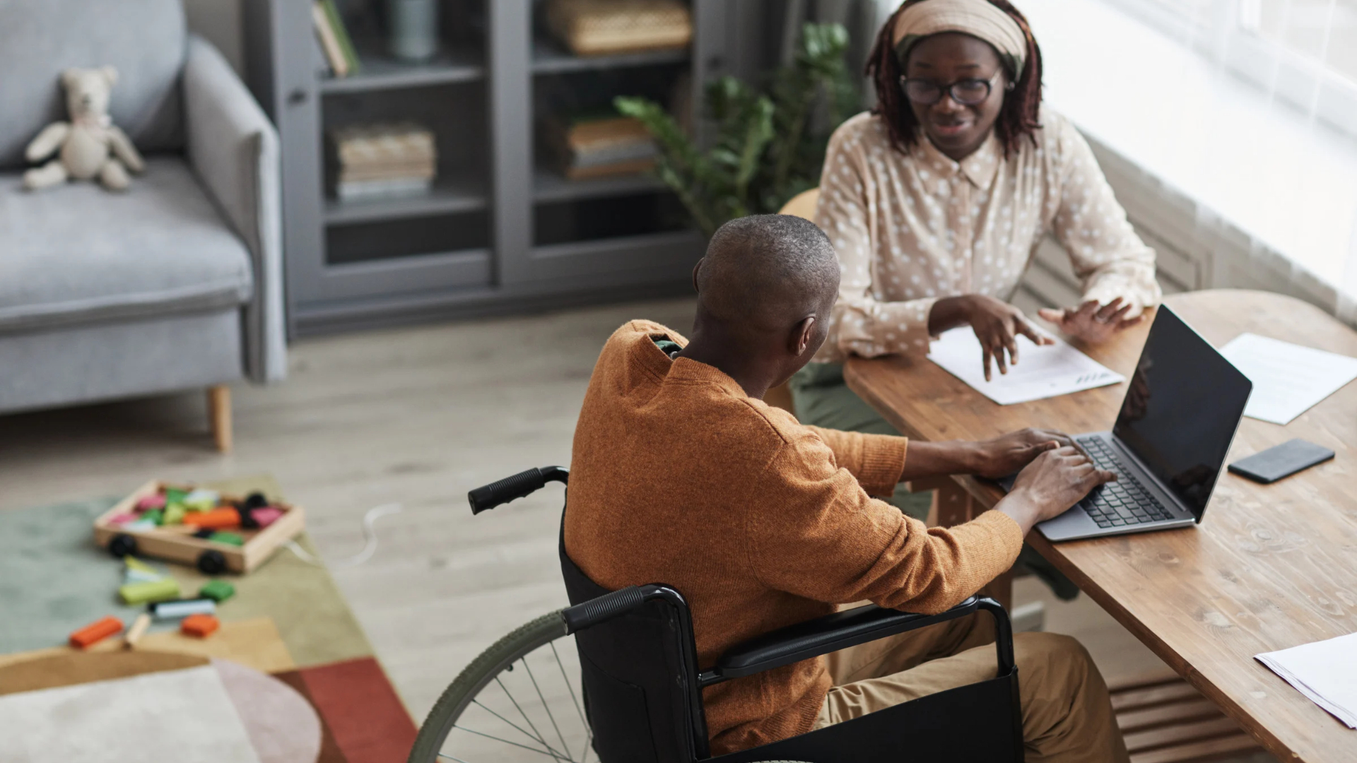 Employee who uses a wheelchair works on a laptop at home while another person reviews papers nearby