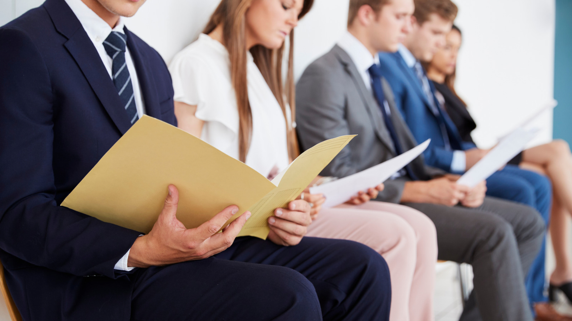 Row of job applicants holding resumes and folders while waiting to interview for the same position.