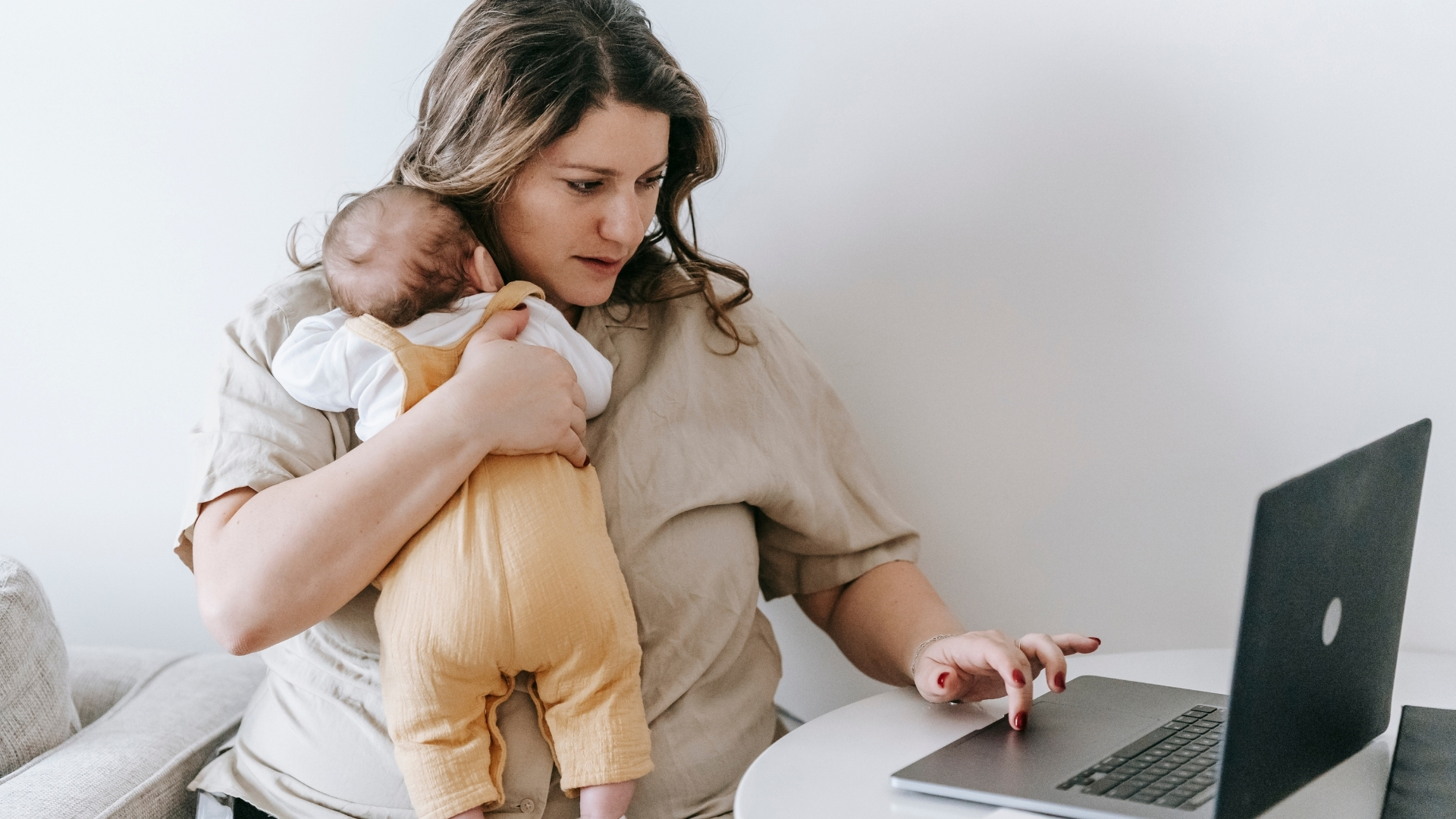 A new mother holds her baby while working on a laptop at home during parental leave.