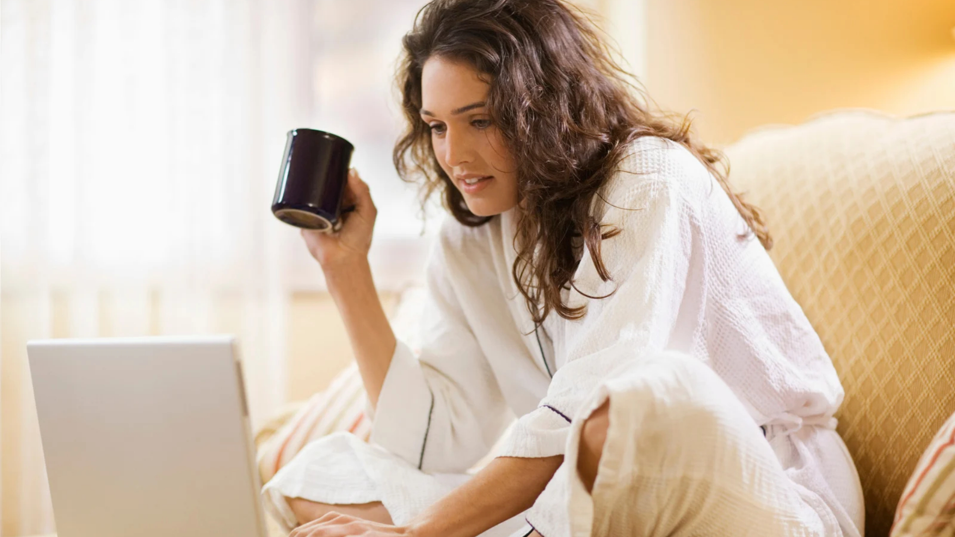 Woman in a bathrobe checking her laptop on the couch at home, holding a coffee mug