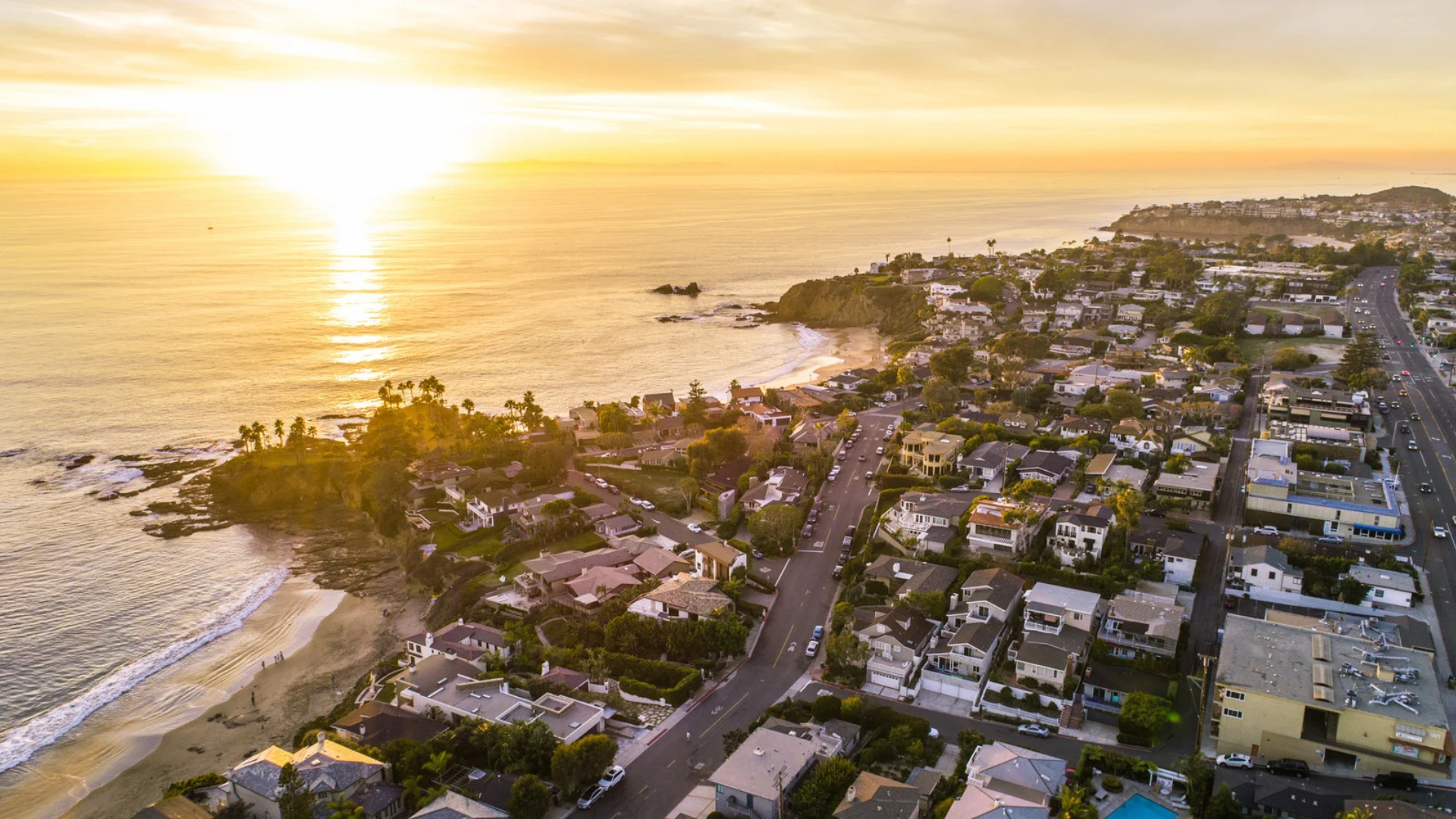 Aerial view of an Orange County coastal neighborhood at sunset with palm trees and homes above the beach